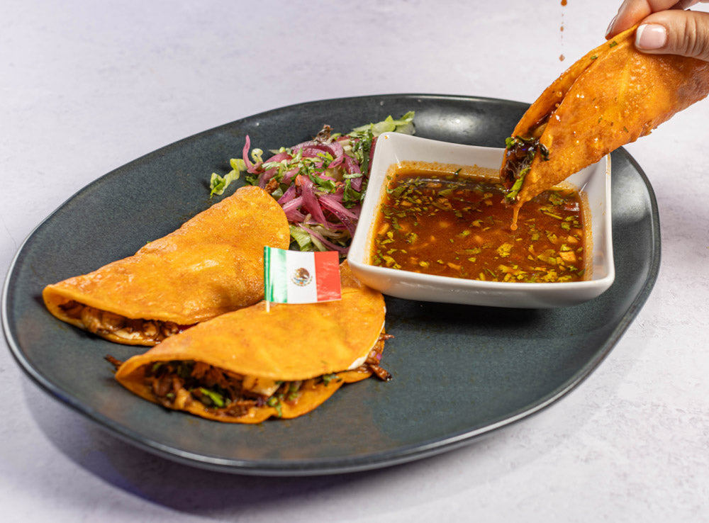 2 Tacos de birria over a black plate, next to a hand holding a juicy birria taco dipped in a a birria sauce.